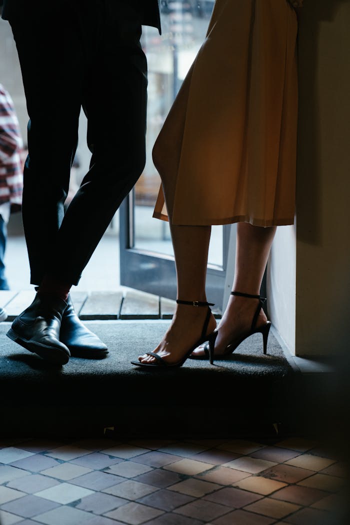 A couple stands elegantly at a doorway, showcasing stylish footwear in a dimly lit setting.